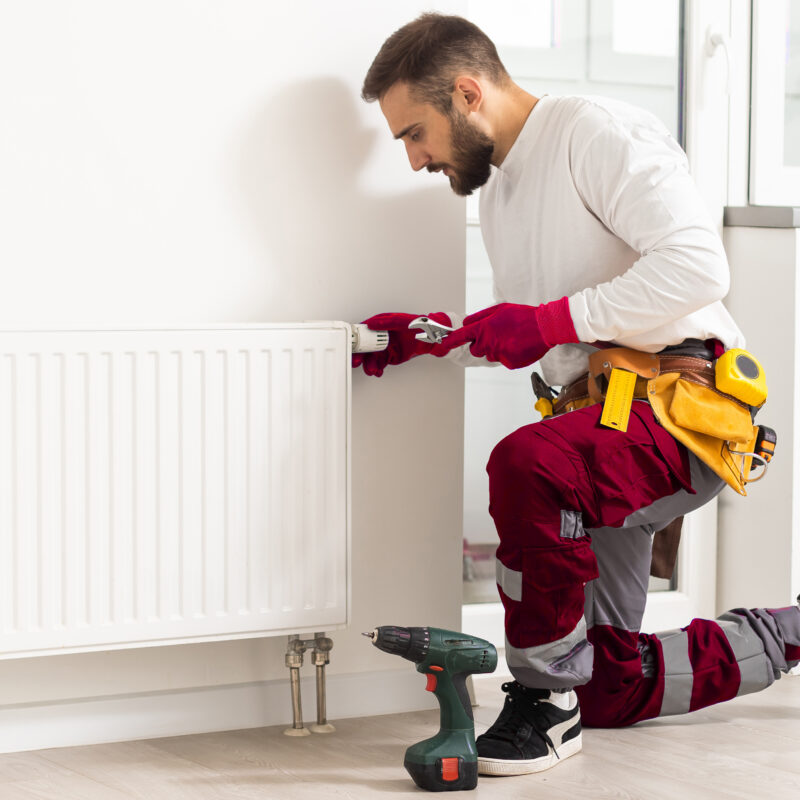 Man in work overalls using wrench while installing heating radiator in room. Young plumber installing heating system in apartment. Concept of radiator installation, plumbing works and home renovation.